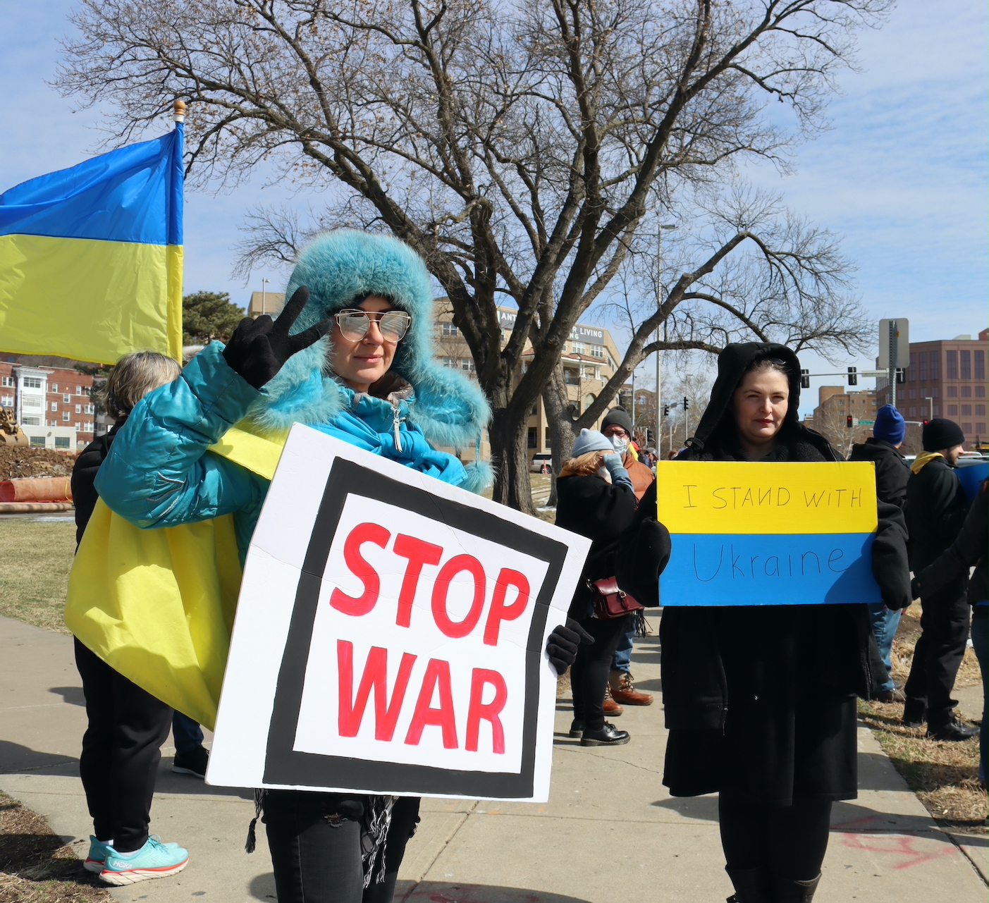 Woman in blue had holds sign that reads "stop war" she also holds up two fingers in a peace salute. Other woman on the right holds a yellow and blue sign that reads "I stand with Ukraine."