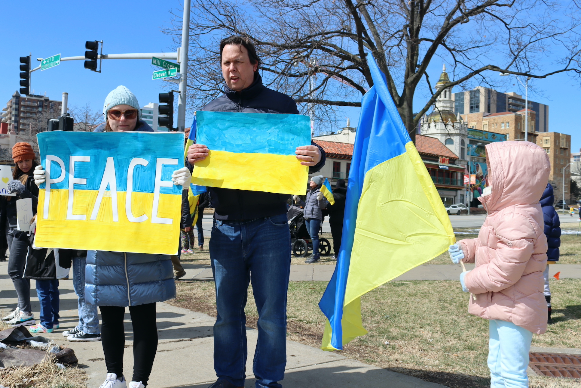 Woman hols poster that reads "peace" next to tall man holding a poster of Ukraine's flag. A young girl in a pink coat holds a Ukrainian flag on a pole.