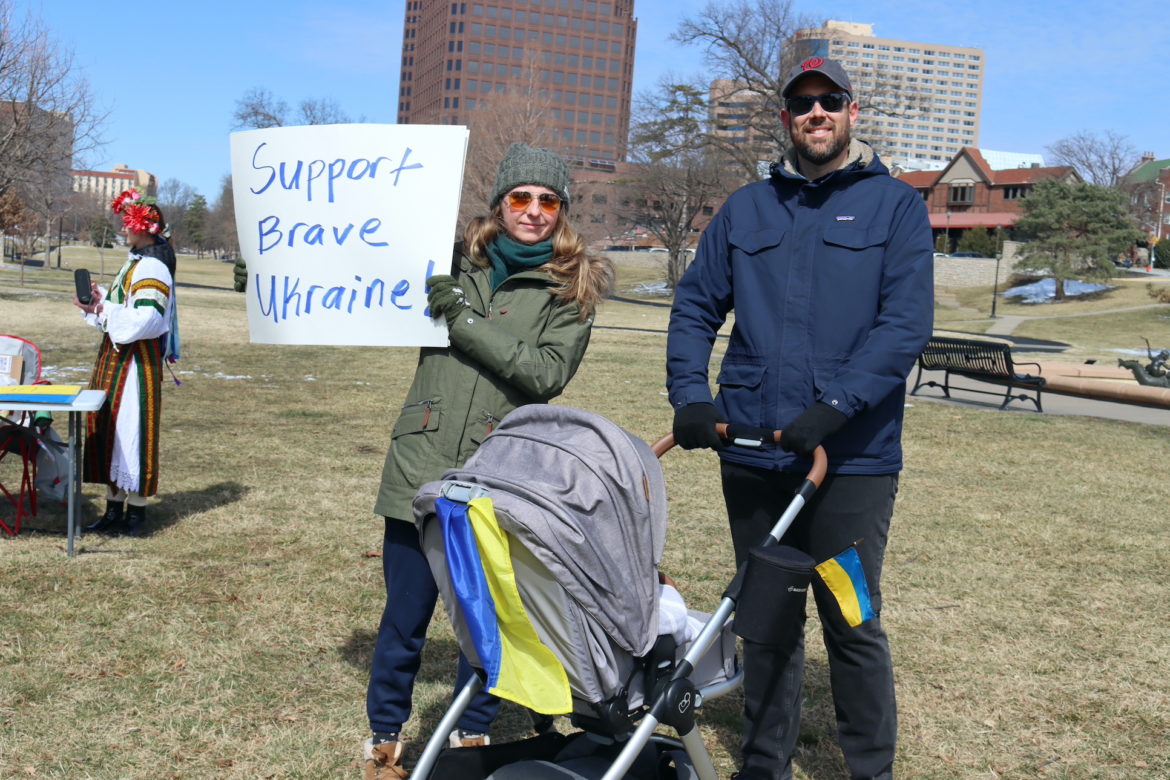 Woman holds a sign that reads "Support Brave Ukraine" next to a man in blue coat pushing a baby stroller covered in Ukrainian flags.