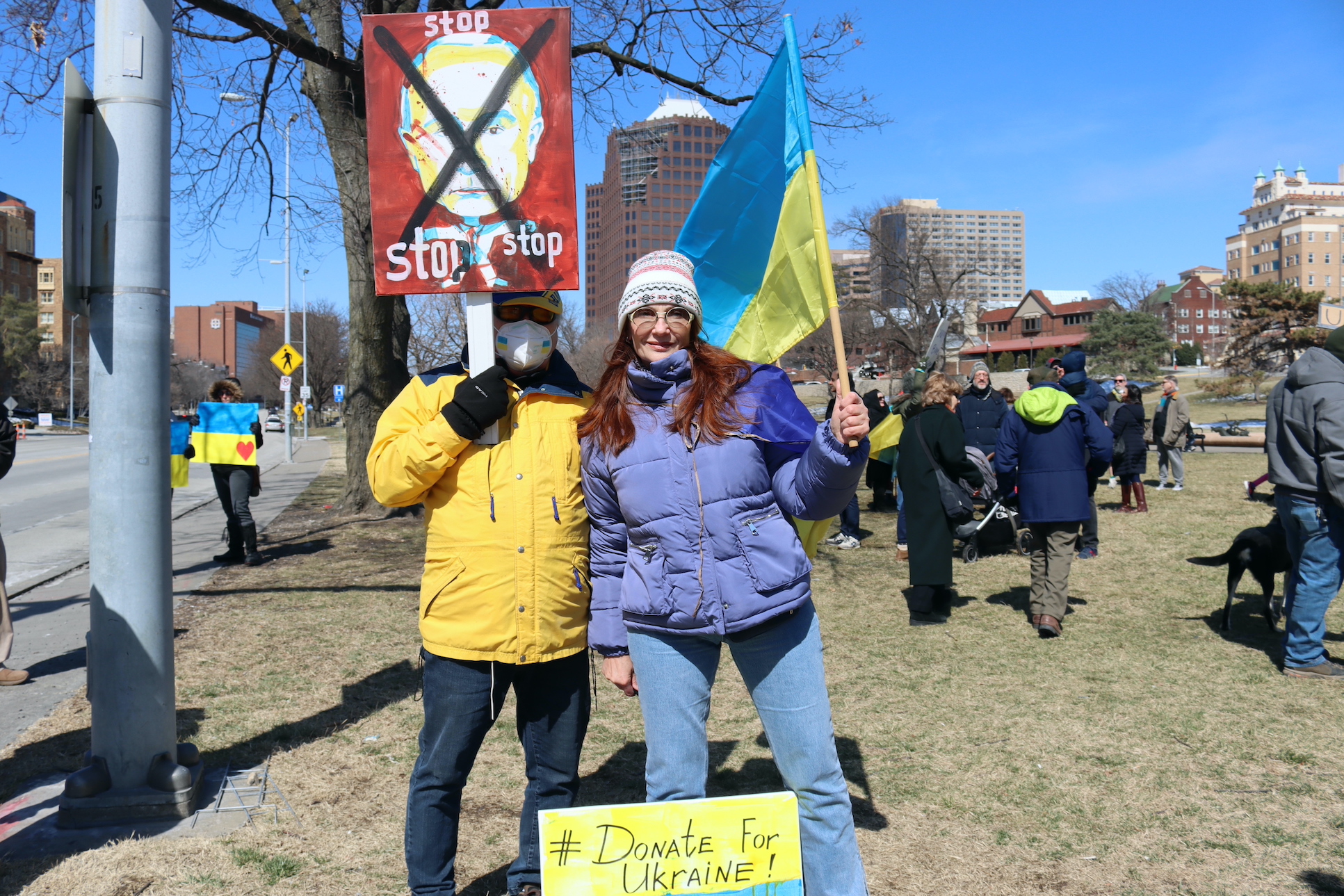 Man holds a sign that reads "stop" with a painting of Vladimir Putin with a big "x' over his face. Woman stands next to him holding a Ukrainian flag.