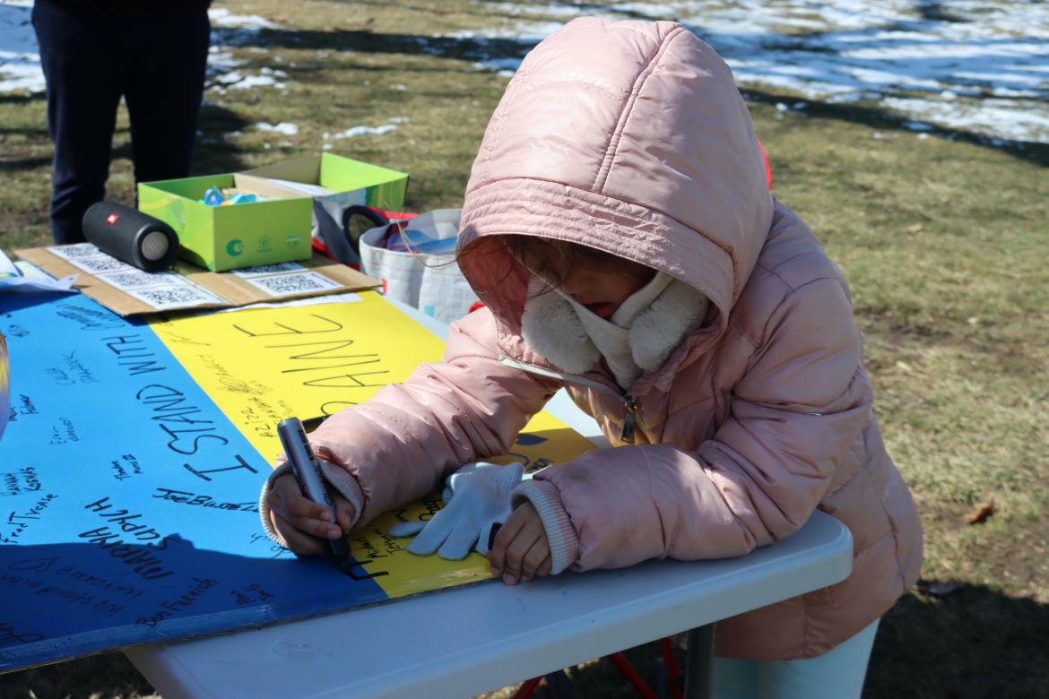 Young girl in pink coat signs her name on a poster of Ukraine's flag. A piece of cardboard shows QR codes behind her.