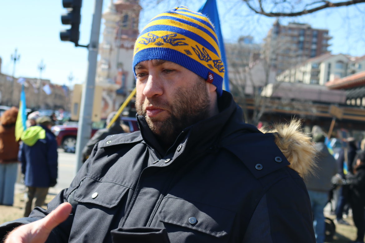Man with blue and gold stocking cap on talks while Ukrainian flags wave in the background in a park.