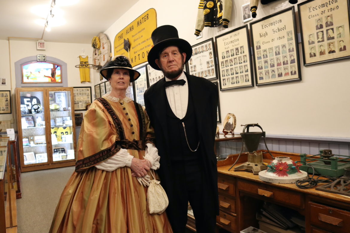 Woman dressed in 19th century gold dress with hat and gloves stands next to man in black suit with top hat.