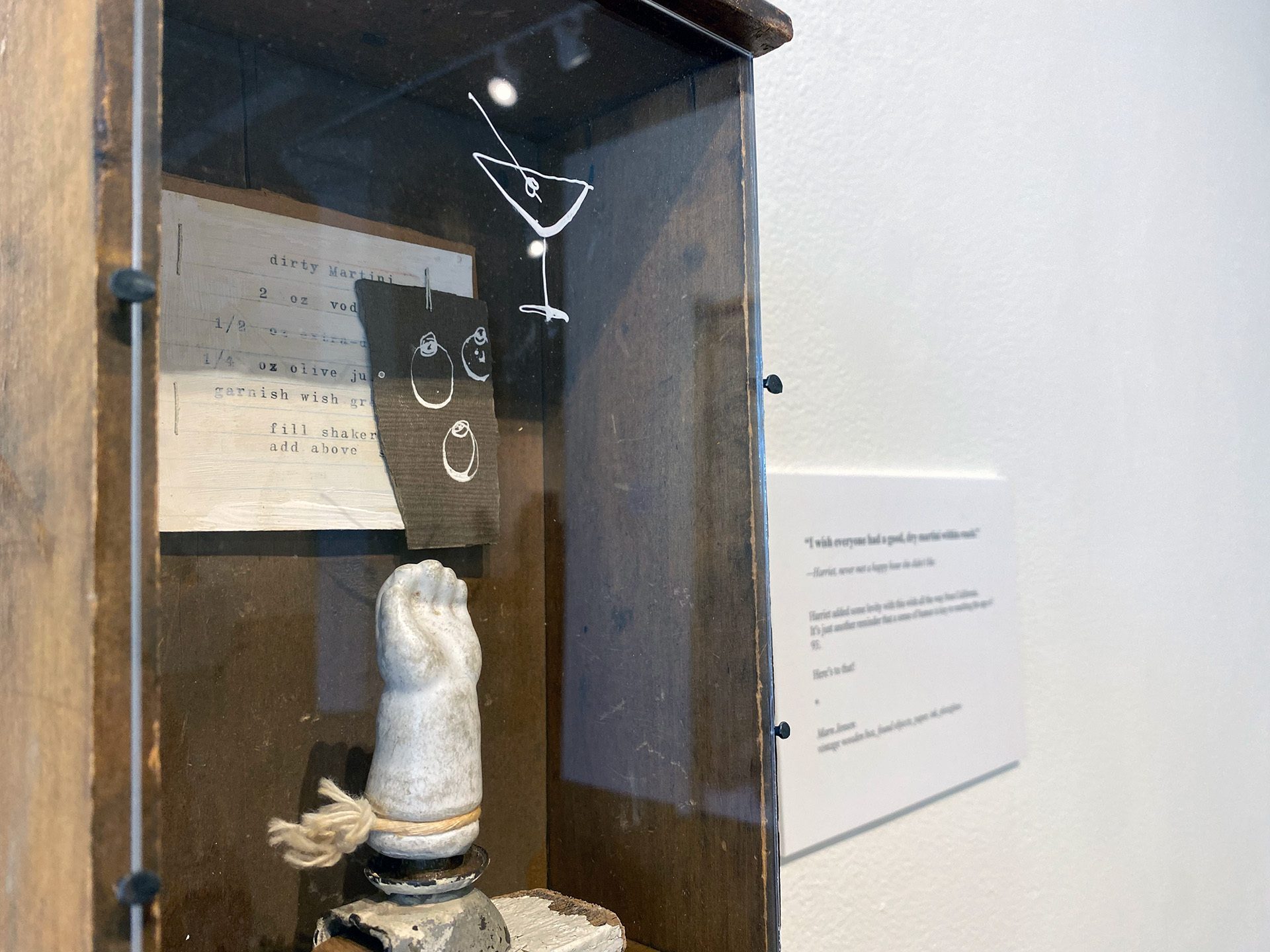 A glass encased porcelain doll hand points upward, reaching toward a white-ink painted martini glass. Some elders had cheeky wishes that centered on having a good time.